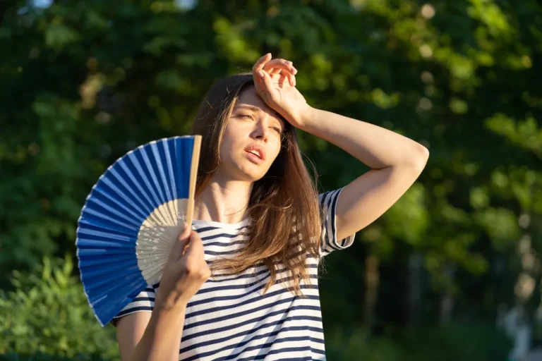 Understanding AC maintenance with MaxCare - Young woman holding a hand held paper fan while wiping sweat from her forehead on a hot summer day.